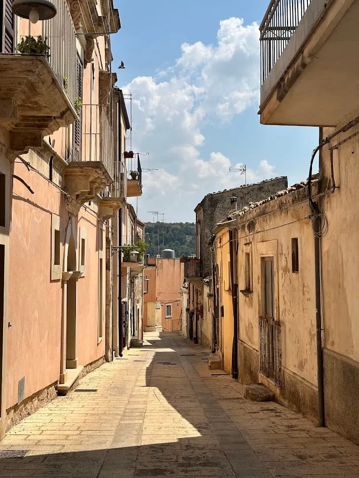 Un callejón soleado con balcones de hierro forjado y una colina verde al fondo en Avola