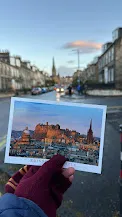 Una postal del castillo de Edimburgo sostenida frente a una calle de la ciudad desenfocada