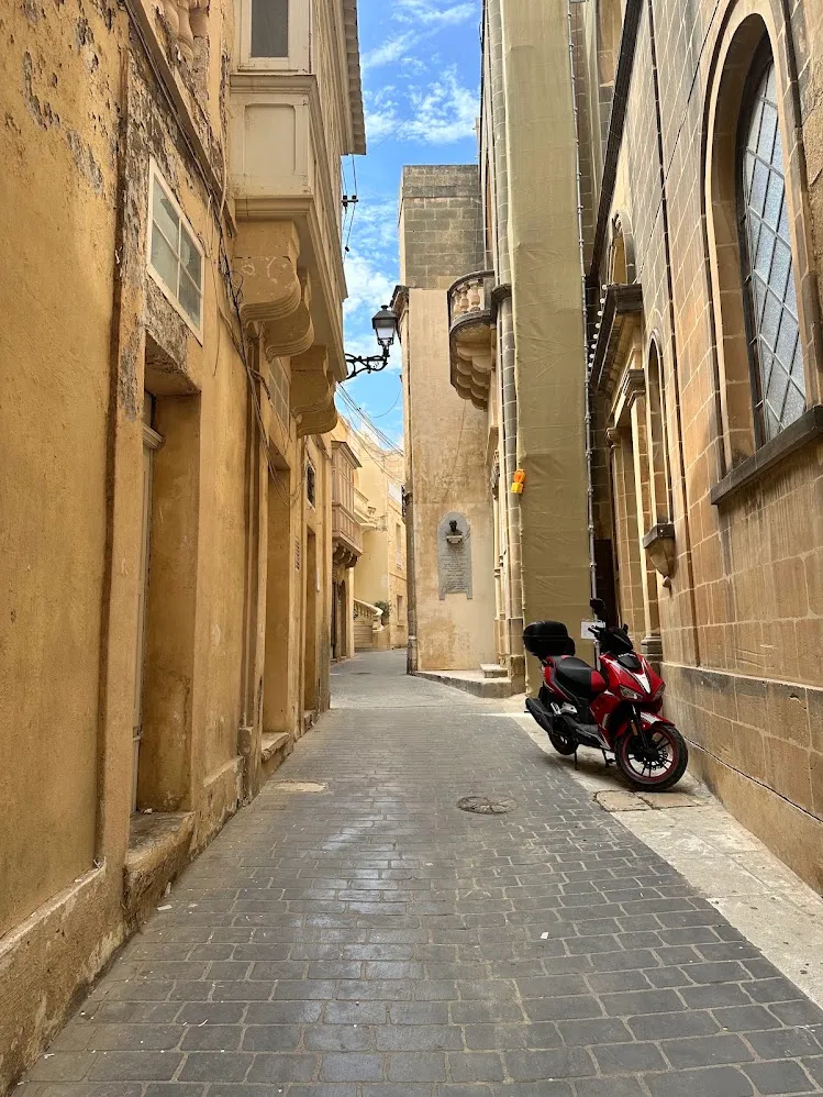 Una calle angosta color miel con una moto roja en Victoria, Gozo